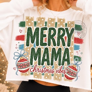 May include: A white long-sleeve sweatshirt featuring a graphic print with the text MERRY MAMA christmas vibes in green and red. The design includes gold vertical stripes with white polka dots, red and blue brush strokes, and two red ornaments.