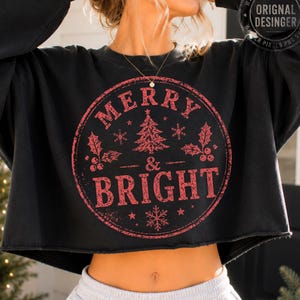 May include: A black long-sleeve cropped sweatshirt featuring a circular red graphic with a distressed texture. The design includes a central Christmas tree, holly leaves, snowflakes, and the text Merry & Bright in a bold, weathered font.