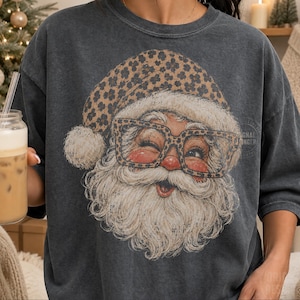 May include: A charcoal gray short-sleeve t-shirt featuring a graphic print of Santa Claus wearing a leopard print hat and matching leopard print glasses. Santa has a white beard, rosy cheeks, and is winking. The shirt has a crew neckline.