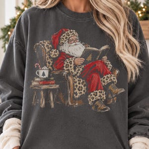 May include: A charcoal grey crewneck sweatshirt featuring a graphic of Santa Claus sitting in a leopard print armchair, reading a book. Beside him is a small table with a mug containing a candy cane and a stack of books.