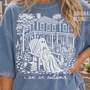 May include: A blue short-sleeve t-shirt featuring a white graphic print of a ghost sitting in a chair on a porch in front of a house. The design includes a stack of books, trees, and the text "i am an autumn" written in a cursive script at the bottom.