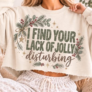 May include: A cream-colored cropped sweatshirt featuring the text I FIND YOUR LACK OF JOLLY disturbing in green and brown lettering. The design is decorated with pine branches, red berries, a bow, and small gold stars around the text.