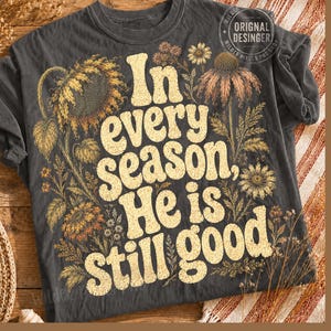 Puede incluir: Una camiseta gris carbón que presenta un gráfico grande de color crema con el texto "In every season, He is still good." El diseño está rodeado de ilustraciones detalladas de girasoles, margaritas y flores silvestres en tonos amarillos y marrones.