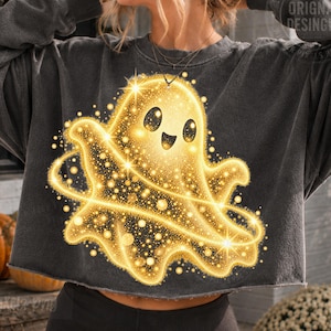 May include: A charcoal grey cropped sweatshirt featuring a large, glowing golden ghost graphic. The ghost has a cheerful expression with black eyes and a wide smile, surrounded by shimmering golden sparkles and a ring of light encircling its body.