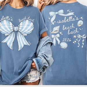 May include: Blue t-shirt with a large bow graphic in light blue and white stripes. The text "isabella's beach bach" is above the bow. The back of the shirt features the same text and bow, along with seashells and the year 2026.
