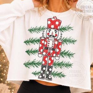 May include: A white long-sleeve sweatshirt featuring a graphic print of a red nutcracker with white polka dots. The nutcracker is positioned over four horizontal green pine branch patterns. The sweatshirt has a cropped hem and long sleeves.