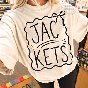 May include: An off-white short-sleeve t-shirt featuring a black graphic print. The design includes a wavy-edged rectangular border, a bow at the top right corner, and the text JAC KETS centered in a bold, rounded, hand-lettered style font.