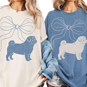 May include: Two long-sleeved shirts, one cream and one blue, each featuring a large bow and a pug graphic. The pug is in a contrasting color to the shirt. The shirts have a relaxed fit and are suitable for casual wear.