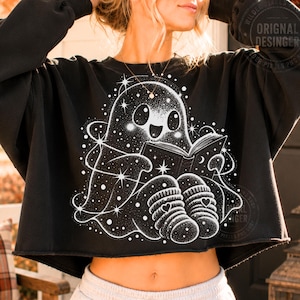 Peut inclure: Un sweat-shirt noir court avec une impression graphique blanche représentant un fantôme lisant un livre. Le fantôme est entouré d'étoiles et de points, et porte des chaussettes rayées avec des cœurs. Le motif a une texture pointillée.