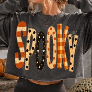 May include: A charcoal gray cropped sweatshirt featuring the word SPOOKY in large, decorative letters. Each letter has a unique pattern: orange and cream stripes, black polka dots, black with orange stars, orange dots, orange plaid, and black polka dots.