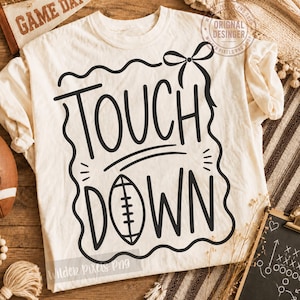 Puede incluir: Una camiseta de manga corta color crema con texto gráfico negro que dice TOUCH DOWN. El diseño incluye una ilustración de un balón de fútbol americano que reemplaza la letra O en DOWN, rodeada por un borde ondulado y un lazo arriba.