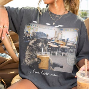 Może przedstawiać: Ciemnoszara koszulka z szopem w okularach przeciwsłonecznych i dżinsowej czapce, pijącym mrożoną kawę ze słomką. Szop znajduje się w klasie. Tekst "i live here now" jest wydrukowany poniżej.