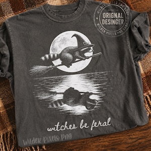 May include: A charcoal gray t-shirt featuring a white graphic of a raccoon wearing a witch hat and riding a broomstick in front of a full moon. The design includes a reflection of the raccoon on water and the text "witches be feral" below.