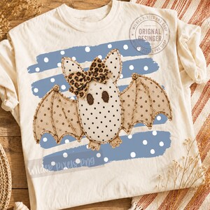 Può includere: Una t-shirt color crema con la grafica di un pipistrello beige a pois neri, che indossa un fiocco con stampa leopardata. Il pipistrello è centrato su uno sfondo a pennellate blu con pois bianchi. Il testo "ORIGINAL DESINGER" è visibile.