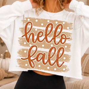 Könnte beinhalten: Ein weißes, langärmeliges, bauchfreies Sweatshirt mit einem Grafikdesign, das den Text hello fall in orangefarbener Schreibschrift zeigt. Das Design umfasst horizontale, beige Pinselstrich-Streifen mit weißen Punkten.