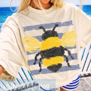 以下が含まれることがあります： クリーム色のTシャツ。手描きの大きなミツバチのデザインが特徴です。ミツバチは黄色と黒で、青い横縞の背景に配置されています。このシャツは半袖で、ゆったりとしたフィット感です。