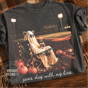 Puede incluir: Una camiseta gris carbón con un gráfico de un fantasma con sábana sentado en una silla plegable en un campo de béisbol por la noche, sosteniendo una taza de café. La escena incluye calabazas, luces y el texto "game day with my boos".