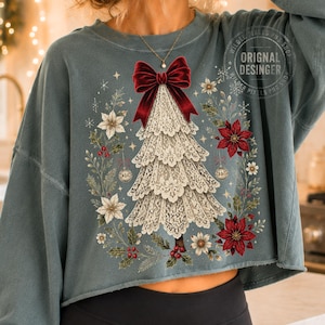 Pu&ograve; includere: Una felpa corta color blu ardesia con la grafica di un albero di Natale in pizzo bianco, sormontato da un grande fiocco in velluto rosso. Il disegno &egrave; circondato da una ghirlanda di stelle di Natale rosse e bianche, fogliame verde e piccoli ornamenti dorati.