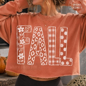 May include: A rust-colored long-sleeve cropped sweatshirt featuring the white text FALL. Each letter has a unique pattern: the F has a floral design, the A has a leopard print, the L has a plaid pattern, and the second L has a polka dot pattern.