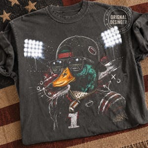 May include: A charcoal grey t-shirt featuring a graphic of a mallard duck wearing a baseball cap and sunglasses. The duck wears a football jersey with the number 1 and a football pendant. The design includes stadium lights and play diagrams.