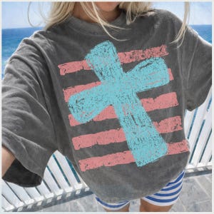 May include: A charcoal gray t-shirt featuring a large, light blue cross over horizontal pink stripes. The design has a distressed, textured appearance. The shirt is part of a casual wear collection.