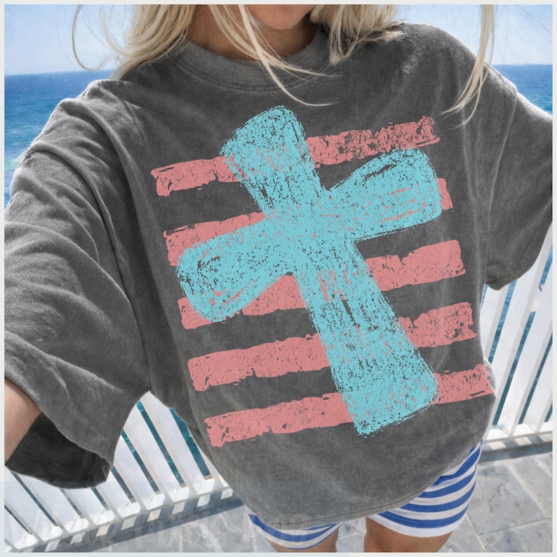 May include: A charcoal gray t-shirt featuring a large, light blue cross over horizontal pink stripes. The design has a distressed, textured appearance. The shirt is part of a casual wear collection.