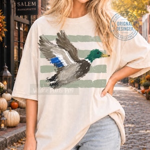 May include: A beige short-sleeve t-shirt featuring a painterly illustration of a mallard duck in flight. The duck has a green head, grey body, and blue wing patch. Behind the duck are three horizontal, distressed sage green stripes.