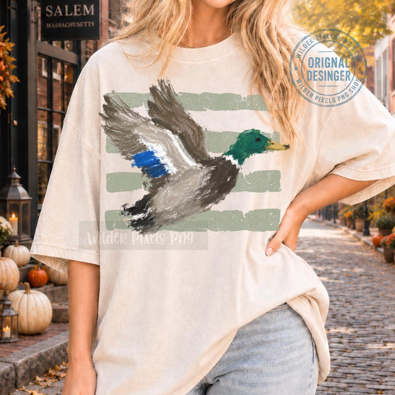 May include: A beige short-sleeve t-shirt featuring a painterly illustration of a mallard duck in flight. The duck has a green head, grey body, and blue wing patch. Behind the duck are three horizontal, distressed sage green stripes.