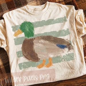 Pu&ograve; includere: T-shirt color crema con un disegno di anatra dipinta, con testa verde, corpo marrone e zampe arancioni. L'anatra &egrave; posta su uno sfondo di strisce orizzontali verdi sbiadite. Il testo "Wilder Pixels Png" &egrave; in basso.