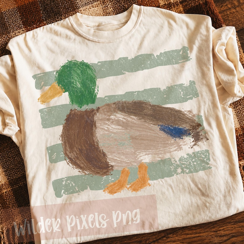 Pu&ograve; includere: T-shirt color crema con un disegno di anatra dipinta, con testa verde, corpo marrone e zampe arancioni. L'anatra &egrave; posta su uno sfondo di strisce orizzontali verdi sbiadite. Il testo "Wilder Pixels Png" &egrave; in basso.