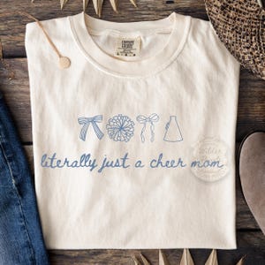 May include: Cream-coloured t-shirt with blue illustrations of a bow, pom-pom, bow, and megaphone. The text "literally just a cheer mom" is written in blue script. The shirt is laid flat on a wooden surface.