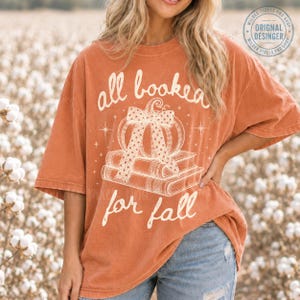 May include: An oversized burnt orange t-shirt featuring a white graphic print of a pumpkin with a polka-dot bow resting on a stack of two books. The text reads all booked for fall in a cursive script, surrounded by small star accents.