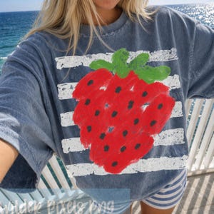 Op de afbeelding: Blauw T-shirt met een grote, handgetekende rode aardbei met zwarte zaadjes en groene bladeren. De aardbei staat tegen een achtergrond van witte horizontale strepen. De persoon draagt het shirt aan zee.
