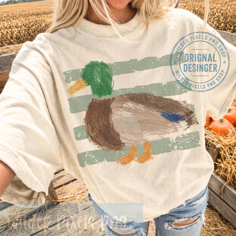 Pu&ograve; includere: T-shirt color crema con un disegno di anatra dipinta con accenti verdi, marroni e blu. L'anatra &egrave; posizionata su strisce orizzontali verde chiaro. La maglietta ha il testo "ORIGINAL DESIGNER" e "WILDER PIXELS PNG SHOP" in un logo circolare.