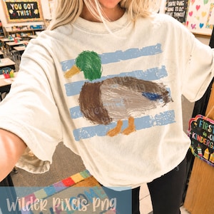 Pu&ograve; includere: T-shirt color crema con un'illustrazione di anatra con testa verde, corpo marrone e zampe arancioni, su strisce orizzontali blu. La maglietta ha una vestibilit&agrave; ampia. Il testo "Wilder Pixels png" &egrave; visibile.