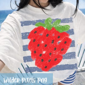 Op de afbeelding: Wit T-shirt met een grote, rode aardbei, groene bladeren en zwarte zaden. Het shirt heeft blauw-witte horizontale strepen en de tekst "Wilder Pixels Png" onderaan.