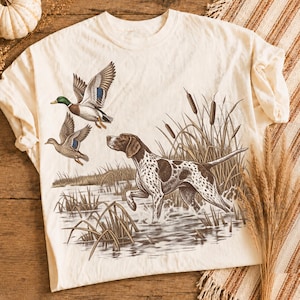 May include: A cream-colored t-shirt featuring a graphic illustration of a brown and white spotted hunting dog standing in water among cattails, with two mallard ducks flying in the upper left corner of the design.