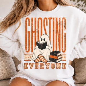 May include: A white crewneck sweatshirt featuring a graphic print with the orange text GHOSTING EVERYONE. The design includes a white ghost character wearing orange and white striped socks while reading a book, next to a stack of three books.