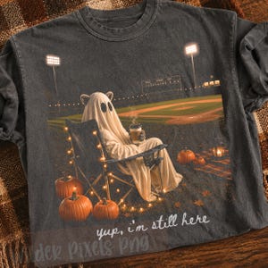 May include: A charcoal gray t-shirt featuring a graphic of a ghost wearing cat ears, sitting in a folding chair on a baseball field at night. The ghost holds a coffee cup, surrounded by pumpkins and string lights. Text reads: yup, i'm still here.