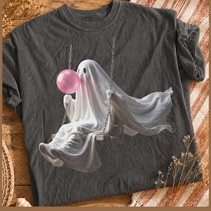 May include: A charcoal gray t-shirt featuring a graphic print of a white ghost sitting on a swing. The ghost is depicted with a round, bright pink bubble gum sphere in front of its face. The fabric of the ghost appears draped and flowing.