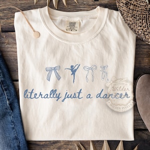 May include: Cream-colored t-shirt with a blue graphic of bows, a ballerina, and ballet shoes. The text "literally just a dancer" is printed in blue script. The shirt is a Comfort Colors brand.