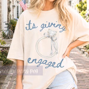 Könnte beinhalten: Cremefarbenes T-Shirt mit dem Schriftzug "its giving engaged" in blauer Schrift. Eine detaillierte Illustration eines Diamant-Verlobungsrings mit Schleife ist mittig auf dem Shirt. Das Shirt hat kurze Ärmel und eine lockere Passform.