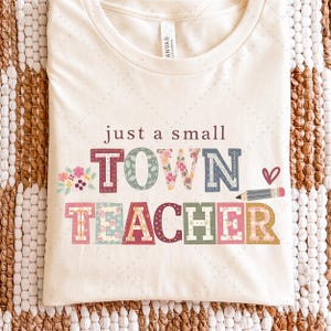 May include: Cream-colored t-shirt with the text "just a small TOWN TEACHER" in colorful, patterned letters. Floral and pencil graphics accent the design. The shirt is folded on a textured, neutral-toned surface.