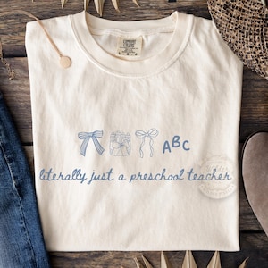 Może przedstawiać: Kremowa koszulka z niebieskim wzorem przedstawiającym kokardy, prezenty i litery "ABC". Tekst "literally just a preschool teacher" jest wydrukowany poniżej. Koszulka jest złożona na drewnianej powierzchni.