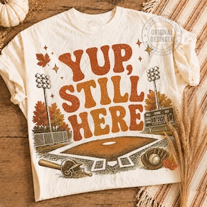 May include: A cream-colored t-shirt featuring a graphic of a baseball field with stadium lights, a scoreboard, a bat, a helmet, and a glove. The text "YUP, STILL HERE" is printed in large, distressed orange and brown letters above the field.