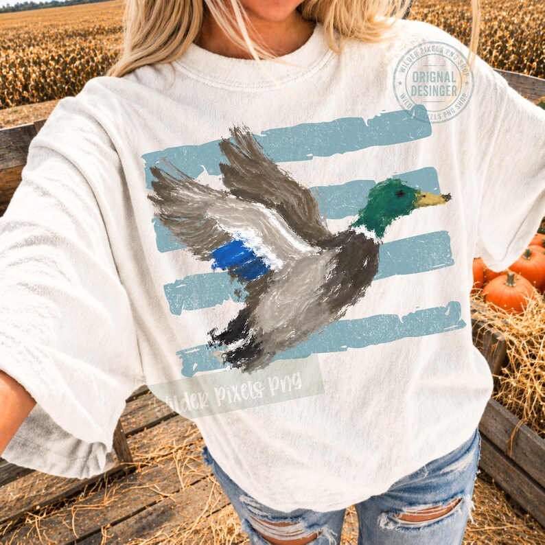 May include: An off-white t-shirt featuring a painterly graphic of a mallard duck in flight. The duck has a green head, yellow beak, and grey wings with a blue patch. Behind the duck are horizontal light blue brushstroke stripes.