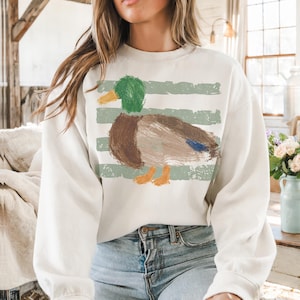 Pu&ograve; includere: Felpa color crema con un design a forma di anatra in stile dipinto a mano. L'anatra ha la testa verde, il corpo marrone e le zampe arancioni. Strisce verdi orizzontali sono dietro l'anatra. La felpa &egrave; uno stile casual e confortevole.