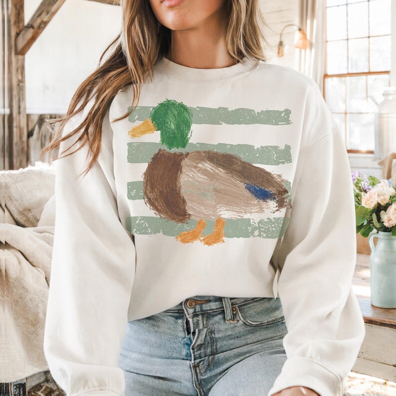 Pu&ograve; includere: Felpa color crema con un design a forma di anatra in stile dipinto a mano. L'anatra ha la testa verde, il corpo marrone e le zampe arancioni. Strisce verdi orizzontali sono dietro l'anatra. La felpa &egrave; uno stile casual e confortevole.