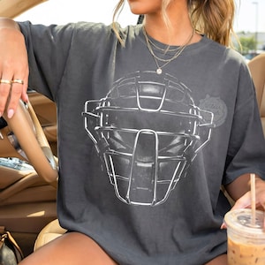 Puede incluir: Camiseta gris oscuro con un gráfico blanco de una máscara de receptor. La camiseta tiene un corte holgado y mangas cortas. El texto "Wilder Roots" está impreso en el lado derecho de la camiseta. La persona lleva anillos y collares dorados.