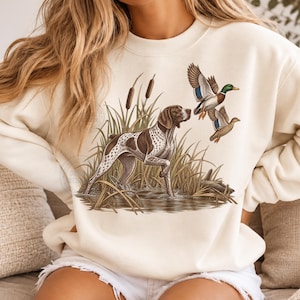 May include: A cream-colored crewneck sweatshirt featuring a graphic illustration of a brown and white spotted hunting dog standing in water among tall grasses and cattails, with two mallard ducks flying above the scene in the center.